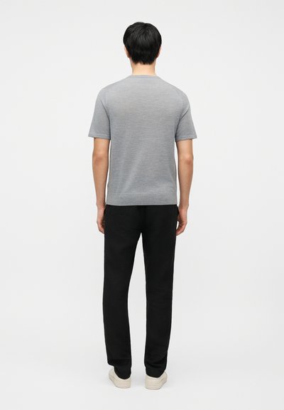 Άνδρας που στέκεται πλάτη προς τα εμπρός, φορά κοντομάνικο γκρι πουκάμισο, μαύρο παντελόνι και λευκά αθλητικά παπούτσια σε απλό λευκό φόντο.