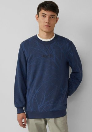Jonge man in een marineblauwe sweatshirt met een donker takkenpatroon en beige broek, staand tegen een effen lichte achtergrond.
