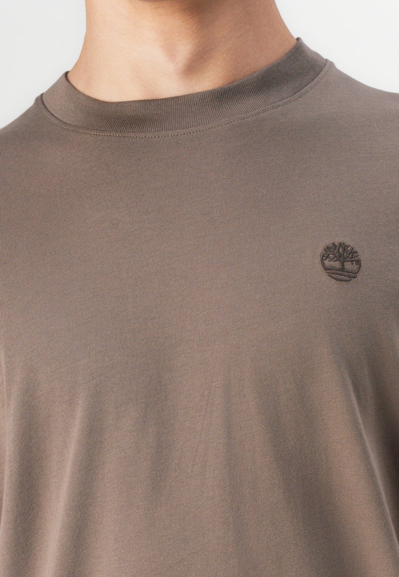 T-shirt à manches courtes, col rond, de couleur brun discrète, fabriqué en tissu doux. Présente un petit logo d'arbre en relief sur la poitrine.