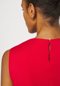 Röd ärmlös klädesplagg med rundad halsringning och en liten nyckelhålsspringa på ryggen, tillverkat av slätt tyg med matt yta.