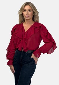 Czerwona, przezroczysta bluzka z warstwowymi falbanami, dekoltem w serek i szerokimi, zgromadzonymi rękawami. Połączona z ciemnoniebieskimi dżinsami z wysokim stanem i paskiem.