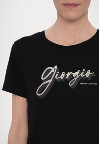 Mustmustunud puuvillane T-särk, millel on suur, läikiv "Giorgio" peegeldavas kirjas ja väiksemas fontis alltekst "ARMANI EXCHANGE".