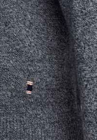 Grauer Strickpullover-Stoff mit einem strukturierten Muster, versehen mit einem kleinen Stoffetikett mit roten und marineblauen Akzenten nahe dem Saum.