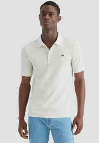 Camisa polo de algodón en tono beige claro, con un tapeta de tres botones, mangas cortas y un pequeño logo bordado en el pecho. Combinada con jeans azules.
