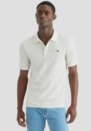 Camisa polo de algodón en tono beige claro, con un tapeta de tres botones, mangas cortas y un pequeño logo bordado en el pecho. Combinada con jeans azules.