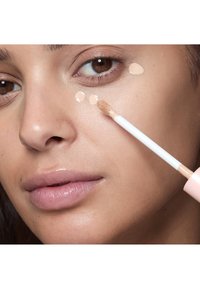 Femme appliquant un correcteur liquide beige avec un applicateur sur la zone sous les yeux, peau nette en gros plan.