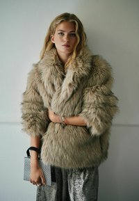 Faux fur frakke i lys beige med voluminøse ærmer, parret med en metallisk sølvfarvet nederdel og holder en lille sølvfarvet clutch. 
