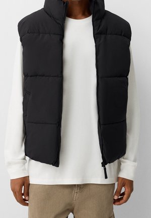 Zwarte doorgestikte mouwloze bodywarmer, open gedragen over een wit shirt met lange mouwen en beige broek, gedragen door een persoon die tegen een effen achtergrond staat.