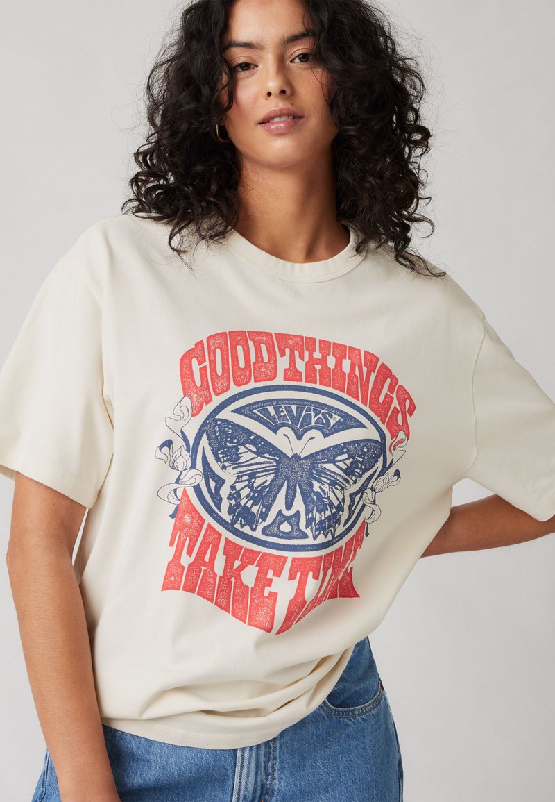 T-shirt oversize couleur crème avec un graphique de papillon bleu et un texte rouge "LES BONNES CHOSES PRENNENT DU TEMPS." En coton, manches courtes.