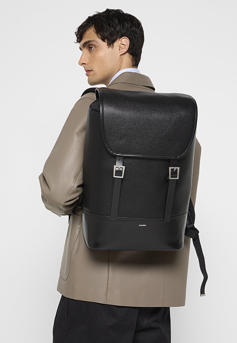 Ein Mann trägt eine taubfarbene Jacke und hat einen großen schwarzen Leder-Rucksack mit Schnallenriemen auf dem Rücken, während er nach links schaut.