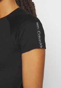 Černá krátká trička s hladkým materiálem, která má elegantní design a potisk loga na levém rukávu.