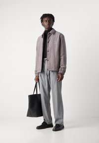 Grå kordfløyelsjakke med elastiske mansjetter, logodetalj; svart turtleneck, rutete bukser og svarte skinnsko. Holder en svart tote bag.