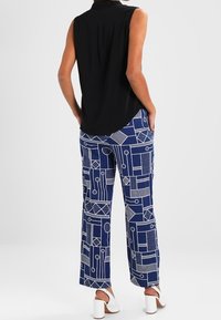 Donna che indossa un blusa nera senza maniche, pantaloni a gamba larga blu e bianchi con motivo geometrico e sandali bianchi con tacco a blocco, in piedi con le mani nelle tasche.