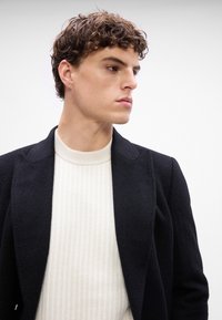 Blazer nero strutturato sopra un maglione bianco a coste, che mostra un design su misura con rever appuntito e dettagli di cucitura sottili.