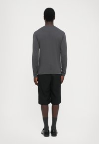 Man wearing a dark gray long-sleeve shirt, black knee-length shorts, dark socks, and black shoes standing facing away on a plain white background.