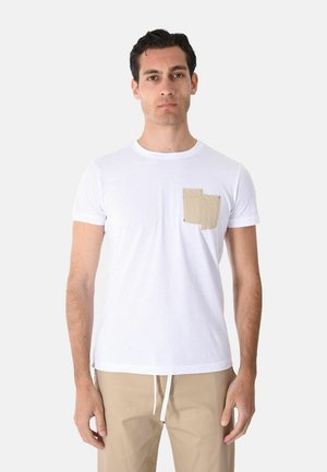 Uomo che indossa una t-shirt bianca a maniche corte con doppia tasca beige, abbinata a pantaloni beige con coulisse, in piedi davanti a uno sfondo semplice.