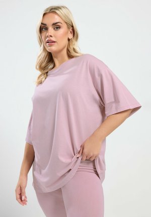 Mujer rubia con camiseta rosa clara de manga corta y suelta y leggings ajustados a juego, sujetando un lateral de la camiseta con una mano.