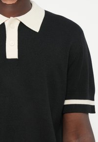 Fekete pólóing, krém színű gallérral és csíkozott mandzsettákkal. Kívül textúrált anyagú, a nyaknál három gombbal. Modern, testhez simuló kialakítás.