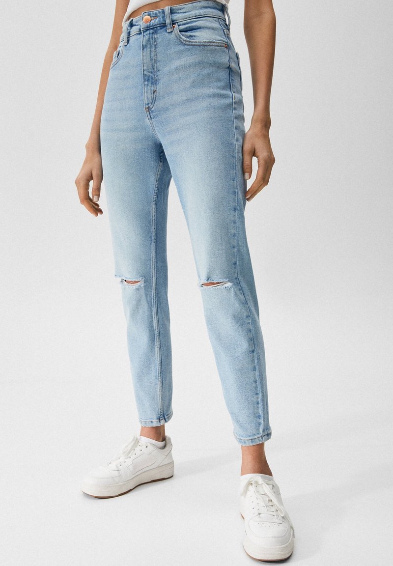 Jeans de tiro alto de color azul claro con detalles desgastados en las rodillas y un corte ajustado. Combinados con zapatillas blancas, mostrando un estilo casual.