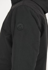 Zwarte winterjas van een gestructureerde stof, met een ronde zwarte logo patch op de linkermouw en een capuchon.