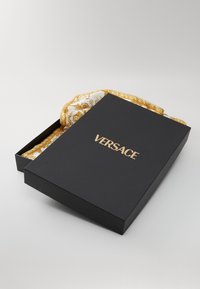 Black rectangular box with gold "VERSACE" lettering, partially open to reveal a patterned silk scarf in white and gold.