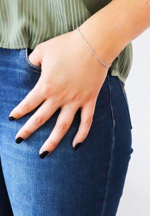 Zilveren kettingarmband gedragen om een hand die in een zak rust, met een textuur, lichtgroen topje en donkerblauwe spijkerbroek. Zwart gelakte nagels zichtbaar.
