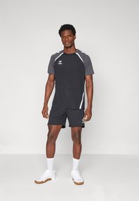 Hombre de pie vestido con ropa deportiva negra y gris, con zapatillas y calcetas blancas, frente a un fondo claro y liso.