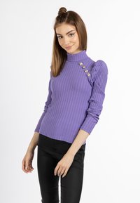 Lila gerippter Rollkragenpullover mit Puffärmeln, versehen mit dekorativen Knöpfen auf einer Schulter. Kombiniert mit schwarzen, eng anliegenden Hosen.
