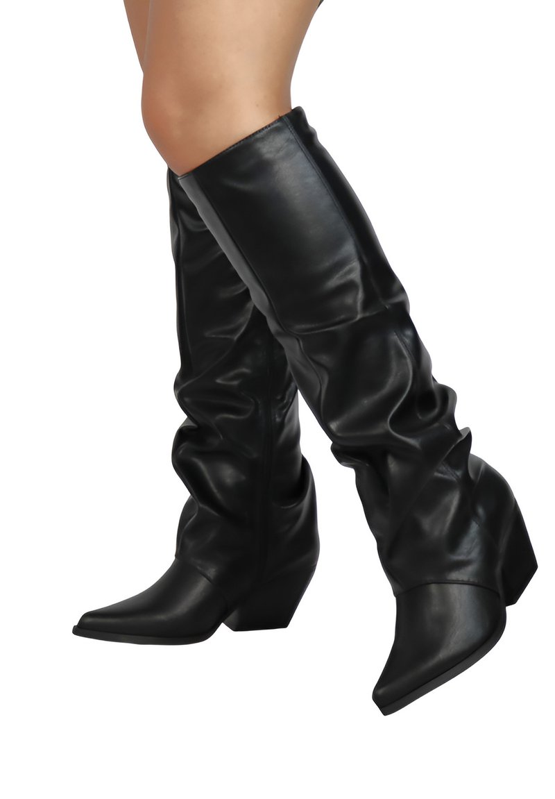 Nogi w czarnych, luźnych, skórzanych kozaczkach do kolan z ostro zakończonymi noskami i blokowym obcasem, przedstawione na białym tle.