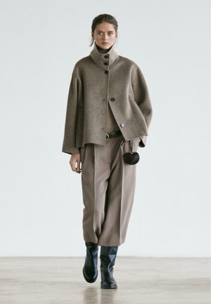 Brown wool blend coat with high collar, button closures, and large pockets; beige knit top, light brown wide-leg trousers, and black boots.