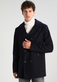 Cappotto peacoat in lana blu marino con ampi rever a nocca, chiusura a doppio petto con bottoni e una vestibilità slim, indossato sopra un dolcevita bianco.