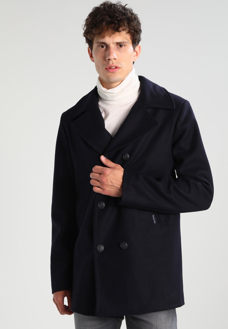 Cappotto peacoat in lana blu marino con ampi rever a nocca, chiusura a doppio petto con bottoni e una vestibilità slim, indossato sopra un dolcevita bianco.