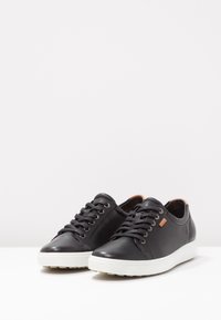 Baskets en cuir noir avec une finition lisse, semelles en caoutchouc blanches, bout rond, laçage marqué et un petit accent beige sur le talon.