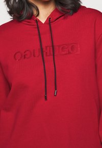 Sweat à capuche en coton rouge avec un logo frontal imprimé ton sur ton et des cordons noirs. Présente un design décontracté et une texture douce.
