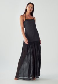 Schwarzes Maxi-Kleid mit einem taillierten Oberteil, dünnen Trägern und einem plissierten, fließenden Rock. Die transparente Textur ergänzt das glatte Oberteil.