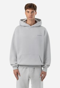 Sweatshirt cinza claro feito de tecido suave, com um bolso frontal, capuz com cordão e uma marca discreta no peito. Corte solto com punhos canelados.