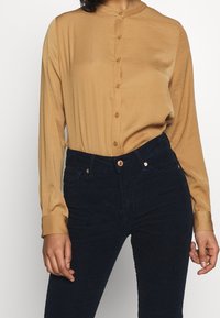 Tan long-sleeve button-up shirt with a round collar and a smooth texture, paired with dark corduroy pants featuring front pockets.
