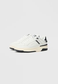 Λευκά αθλητικά παπούτσια με μαύρες λεπτομέρειες, κατασκευασμένα από δέρμα και συνθετικά υλικά. Σχεδιασμός χαμηλού μποτάκι, λαστιχένια σόλα και εμφανές σύστημα κορδονιών.