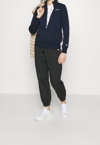 Sudadera con cremallera color navy y mangas largas, joggers negros ajustados y zapatillas blancas. La sudadera tiene un bolsillo en la parte delantera y detalles con el logotipo.