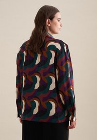 Blusa multicolore con motivo a onde in verde, bordeaux e crema. Maniche lunghe con polsini bottonati, tessuto morbido e vestibilità comoda.