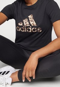 Svart bomulls-T-shirt med ett Adidas-logotyp i leopardmönster, kombinerad med svarta leggings och vita sportskor.
