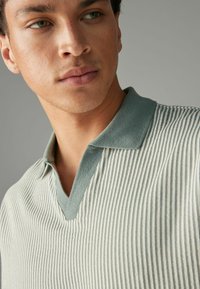 Poloko s dizajnom v-neck; obsahuje vertikálne pruhy v svetlozelenej a smotanovej farbe. Mäkká textúra, kontrastný golier a krátke rukávy.