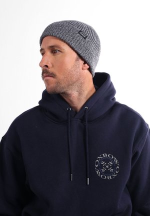 Bonnet en tricot gris avec une texture côtelée, arborant une petite étiquette noire. Porté avec un sweat à capuche bleu marine affichant un logo circulaire.