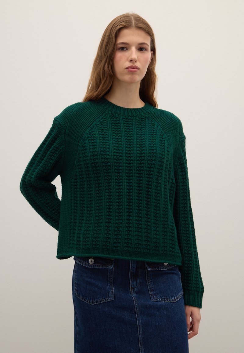 Woman standing against plain background wearing dark green textured knit sweater and blue denim skirt with front pockets.