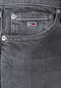 Gray denim jeans with a distressed texture, featuring a small embroidered logo in red, white, and blue on the front pocket. Metal rivets visible.