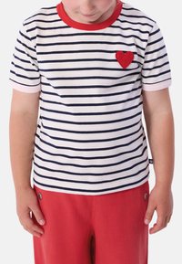 Ριγέ μπλουζάκι με οριζόντιες ναυτικές και λευκές ρίγες, κόκκινη λαιμόκοψη και μικρό κόκκινο τσόχινο καρδιόσημο στο στήθος, συνδυασμένο με κόκκινο παντελόνι.