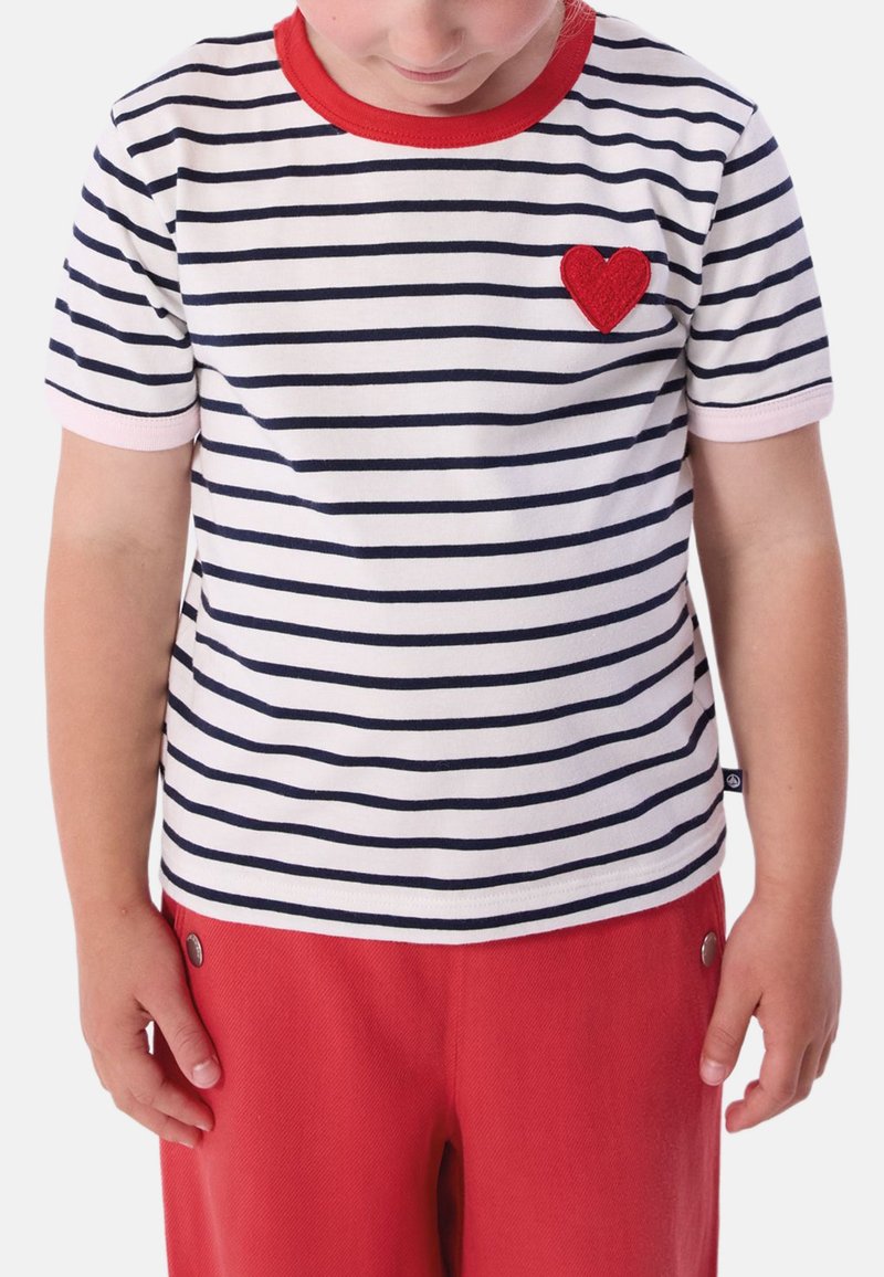Ριγέ μπλουζάκι με οριζόντιες ναυτικές και λευκές ρίγες, κόκκινη λαιμόκοψη και μικρό κόκκινο τσόχινο καρδιόσημο στο στήθος, συνδυασμένο με κόκκινο παντελόνι.