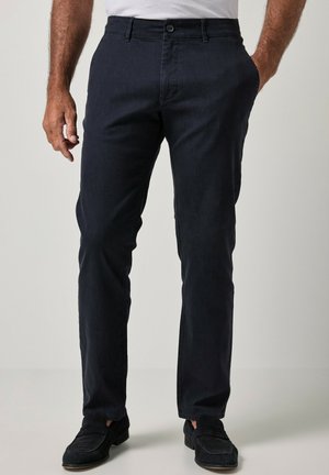 Homme portant un pantalon noir slim-fit, une chemise blanche et des mocassins noirs, debout avec une main dans la poche sur un fond uni.