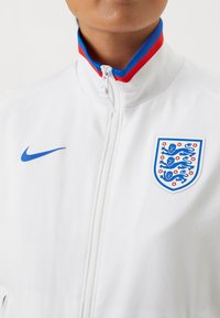 Hvit zip-up jakke laget av glatt stoff, med en blå Nike-logo på venstre bryst og et England-emblem med røde og blå detaljer.
