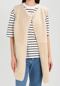 Chaleco de piel sintética beige con diseño de frente abierto, textura suave y sin cierres, llevado sobre una camiseta de rayas blancas y negras y jeans azules.